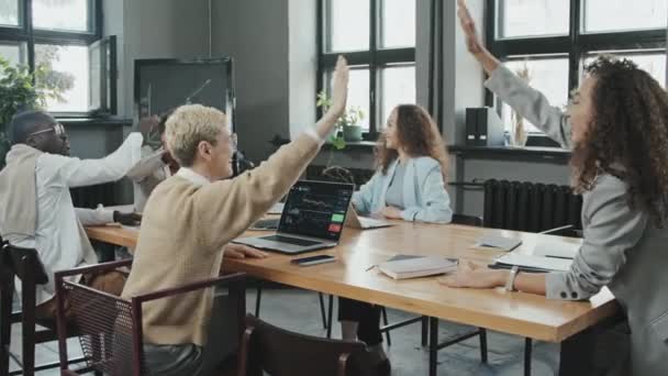Medium slow mo van vrolijke jonge multi-etnische collega 's zitten aan vergadertafel in moderne coworking office, het geven van high-fives aan elk vieren van succesvolle investeringen - Video