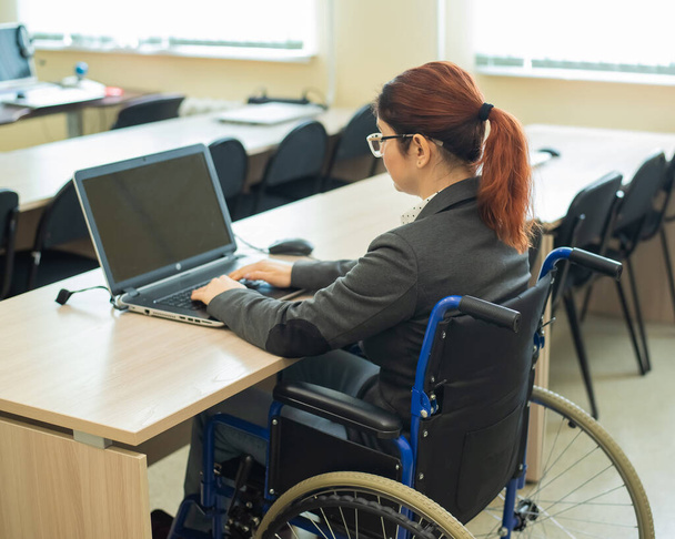 Young woman works at a laptop while sitting in a wheelchair in a university lecture hall. Conditions for teaching a disabled person - Photo, Image