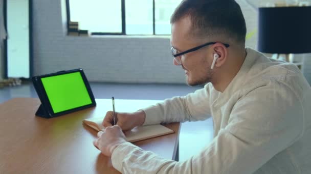 Homem de negócios caucasiano está tomando notas no livro durante uma reunião de vídeo em seu tablet de casa - Filmagem, Vídeo