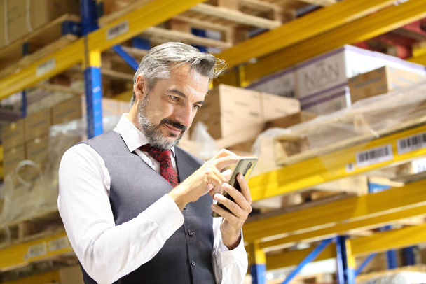 Senior store manager in formal wear using mobile phone to tracking shipment online while checking inventory in distribution warehouse - Photo, image