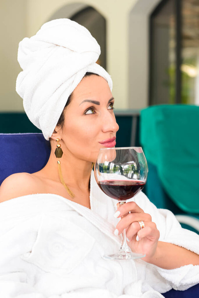 Woman enjoying a glass of red wine in outdoor restaurant. Young caucasian girl in a bathrobe and a towel relaxing with a glass of wine. Fashion, leisure time, girl time. - Foto, afbeelding