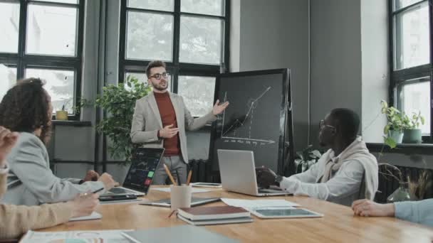 Zoom-out slowmo van zelfverzekerde jongeman voor collega 's die bedrijfspresentatie wijzend naar tekening van grafiek op schoolbord - Video