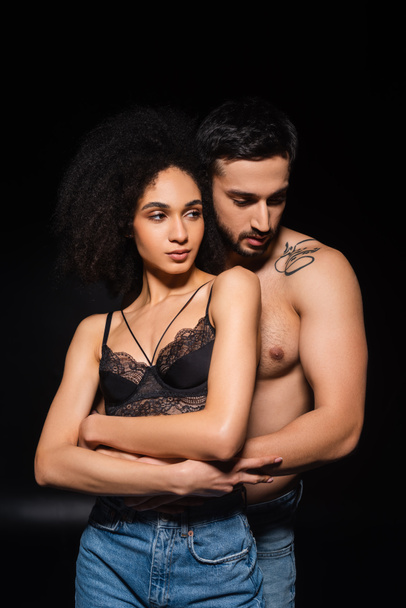 Muscular man embracing african american girlfriend in bra and jeans on black background  - 写真・画像