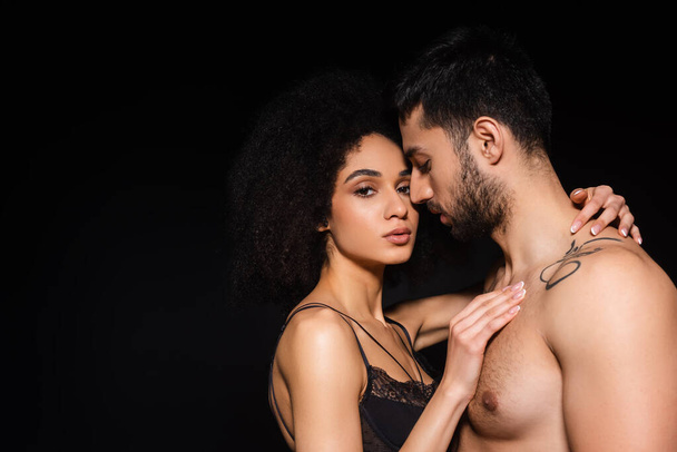 Sexy african american woman looking at camera while embracing shirtless man isolated on black  - Foto, Imagem