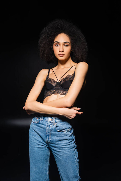 Sensual african american woman in jeans and bra looking at camera on black background  - Foto, Imagen