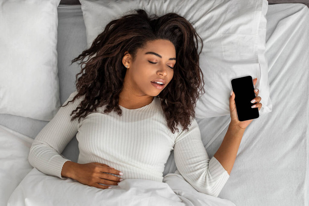 Mujer negra acostada en la cama, durmiendo y sosteniendo el teléfono móvil - Foto, imagen