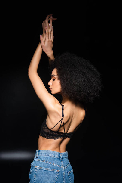Side view of young african american woman in bra and jeans on black background  - Foto, Imagen