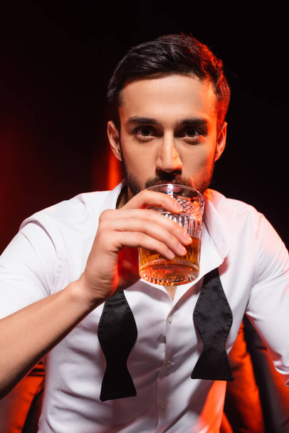 Bearded man in formal wear drinking whiskey on black background with red lighting - Foto, Imagen