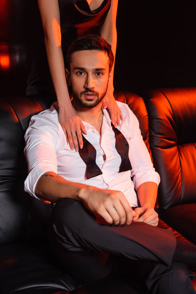 Young woman touching bearded boyfriend on couch on black background with red lighting - Фото, изображение