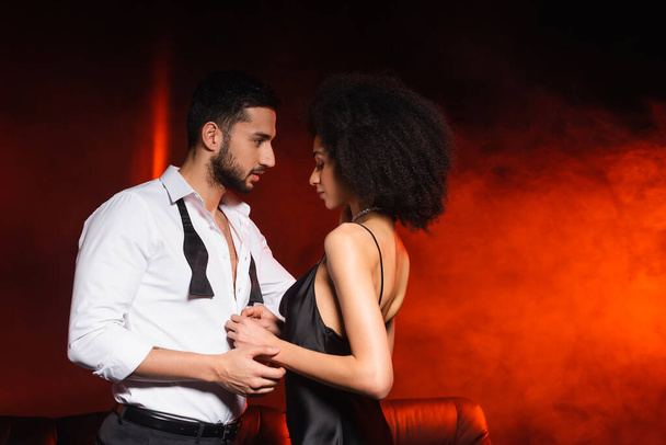 Side view of african american woman in dress standing near boyfriend on black background with red lighting and smoke - Fotó, kép