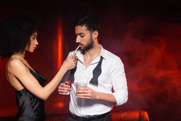 African american woman holding lighter near boyfriend with cigarette and whiskey on black background with red lighting and smoke  - Φωτογραφία, εικόνα