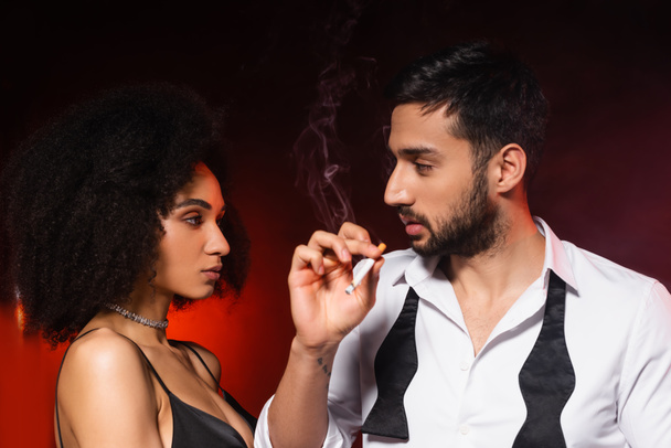 Mann raucht Zigarette in der Nähe eleganter afrikanisch-amerikanischer Freundin auf schwarzem Hintergrund mit rotem Licht - Foto, Bild