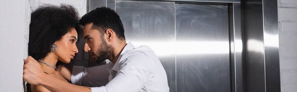 Zijaanzicht van de man met de baard op zoek naar mooie Afrikaanse Amerikaanse vriendin in de buurt van lift, banner  - Foto, afbeelding