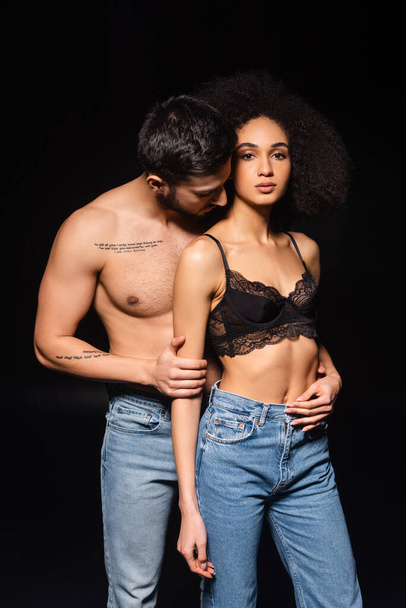 Shirtless man kissing african american woman in jeans and bra isolated on black  - 写真・画像