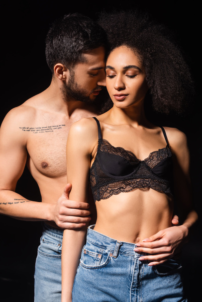Shirtless and bearded man embracing african american woman in bra isolated on black  - Fotoğraf, Görsel
