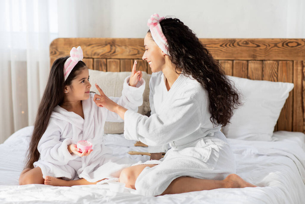 Happy mother and daughter in bathrobes using moisturizer - Photo, Image