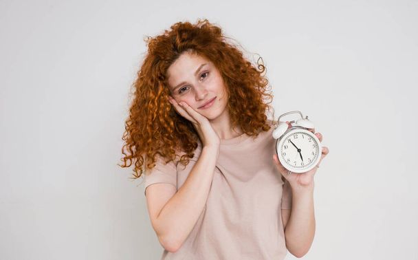 portrait of a beautiful woman with red curly hair holding an alarm clock. Sleep and wake up concept. good morning - Zdjęcie, obraz