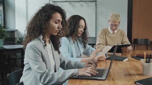 Side-view medium slowmo τριών επιτυχημένων νέων επιχειρηματιών γυναικών που κάθονται σε μεγάλο τραπέζι στο σύγχρονο συνεργαζόμενο γραφείο που εργάζονται σε φορητό υπολογιστή, ψηφιακό tablet που εξετάζει τα έγγραφα των επιχειρήσεων - Πλάνα, βίντεο