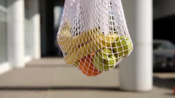 Weiße Netztasche mit Früchten, die sich auf einem Straßenhintergrund drehen. Umweltfreundliche, wiederverwendbare Einkaufstasche. Orangen, Äpfel, Bananen in Baumwoll-Stricktasche. Null Abfall und plastikfreies Konzept. - Filmmaterial, Video