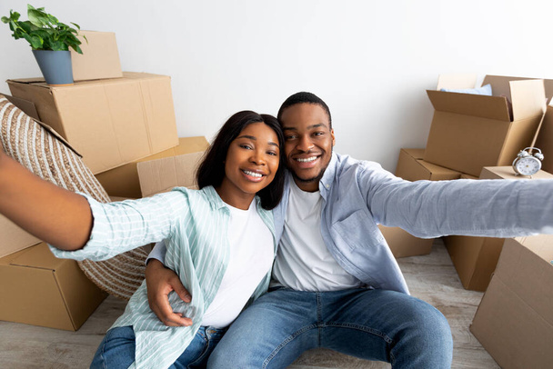 Nadšený africký americký pár dělat selfie po přestěhování do vlastního domu, sedí blízko vybalené krabice a usmívá - Fotografie, Obrázek