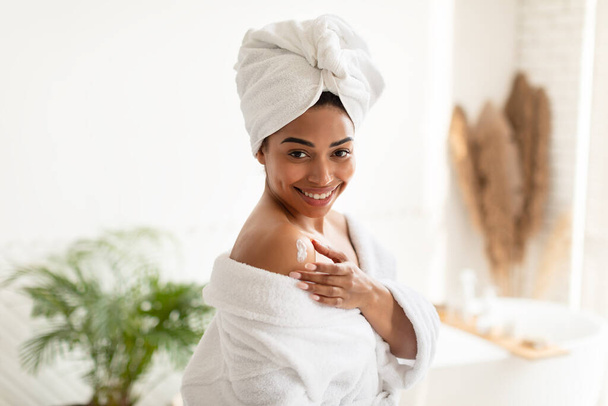 Femme noire appliquant la lotion sur l'épaule debout dans la salle de bain moderne - Photo, image