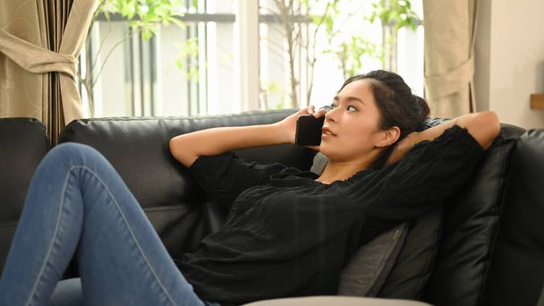 Молода азіатка розмовляє по мобільному телефону, коли лежить на чорному шкіряному дивані.. - Фото, зображення
