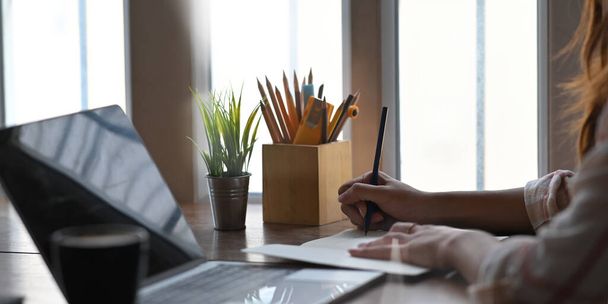 Detailní záběr žena pracující jako básník psaní na notebook, zatímco sedí před počítačem notebook s černou prázdnou obrazovkou na pracovním stole přes pohodlná okna jako pozadí. - Fotografie, Obrázek