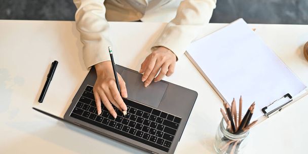 Top view image of businesswoman's hands typing on computer laptop that putting on white working desk and surrounded by pen, pencils in vase and clipboard. Orderly workspace concept. - Foto, imagen