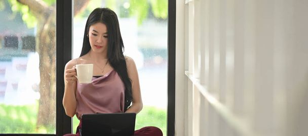 Одна азіатка тримає чашку кави та комп "ютерний лептоп, сидячи на підлозі у вітальні.. - Фото, зображення