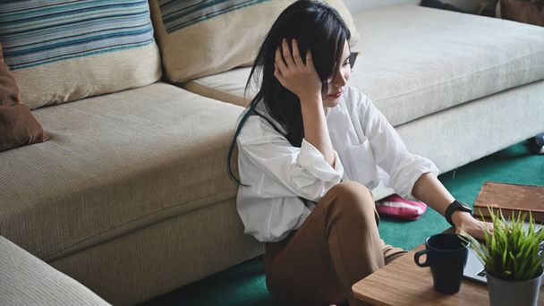 Photo of young beautiful woman relaxing on her holiday/using a computer laptop and enjoying with her coffee while sitting at the comfortable couch over orderly living room as background. - Photo, Image