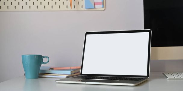 Фото комп'ютерного ноутбука з білим порожнім екраном на білому робочому столі з чашкою кави, стосом книги, персональним комп'ютером та клавіатурою над впорядкованою стіною вітальні як фон
. - Фото, зображення