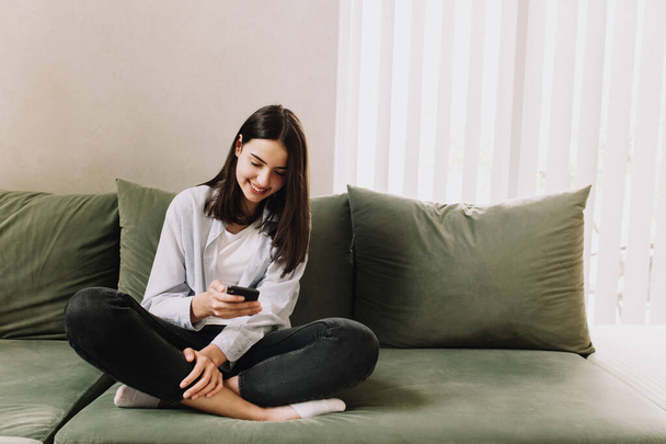 Коммуникационная концепция с черным слышать улыбающуюся девушку, сидящую на зеленом диване, используя и держа в руке смартфон, болтая со своими друзьями - Фото, изображение