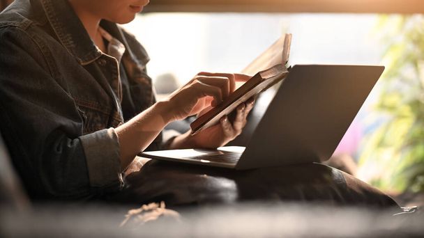 Человек держит и читает книгу, сидя перед компьютером ноутбук на кожаном диване над удобной гостиной в качестве фона. - Фото, изображение