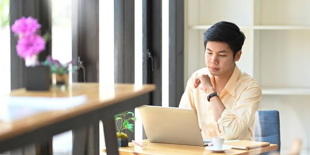 Фото розумного чоловіка, який працює бухгалтером, друкуючи на комп'ютерному ноутбуці, сидячи за дерев'яним робочим столом над комфортною скляною стіною кафе як фон
. - Фото, зображення