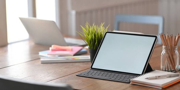 Zdjęcie białych blanków ekranu komputera tablet z klawiaturą na drewnianym biurku z laptopem i sprzętem na wygodnym salonie jako tło. Koncepcja uporządkowanego miejsca pracy. - Zdjęcie, obraz