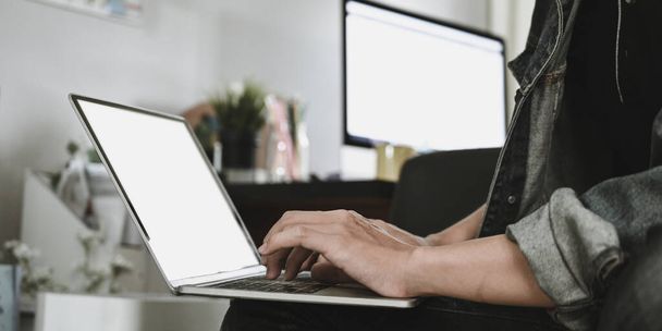 Ausgeschnittenes Bild der Hände des intelligenten Mannes, der auf einem weißen leeren Bildschirm Computer-Laptop tippt, der auf seinem Schoß über dem gemütlichen Wohnzimmer als Hintergrund sitzt. Arbeit von zu Hause aus und Remote-Arbeitskonzept. - Foto, Bild