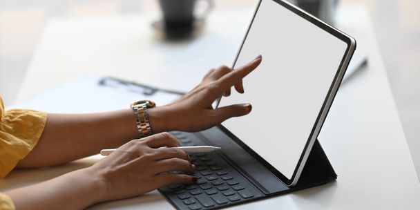 Przycięty obraz rąk kreatywnej kobiety trzymającej długopis i korzystającej z tabletu komputerowego z klawiaturą siedząc przy białym biurku nad wygodnym biurem jako tło. - Zdjęcie, obraz