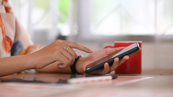 L'immagine ritagliata della mano di una donna utilizza uno smartphone alla scrivania di legno. - Foto, immagini