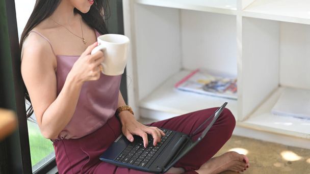 Kesilmiş resimli kadın kahve içiyor ve oturma odasında otururken bilgisayar kullanıyor.. - Fotoğraf, Görsel