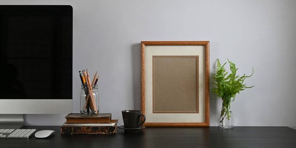 Photo of black working desk along with black blank screen computer monitor, books, notebook, pencil holder, picture frame, potted plant putting together on it with white cement wall as background. - Photo, Image