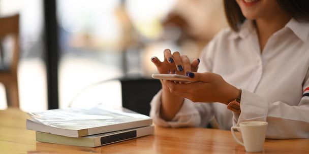Przycięty obraz dłoni młodej kobiety trzymającej smartfona podczas siedzenia przy drewnianym biurku nad wygodnym miejscem pracy jako tłem. Kobieta relaksująca koncepcja czasu. - Zdjęcie, obraz