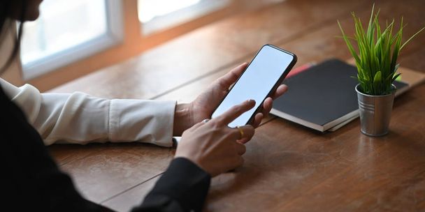 Zugeschnittenes Bild einer stilvollen Frau, die ein weißes Smartphone mit leerem Bildschirm hält und benutzt, während sie am Holzarbeitstisch über dem gemütlichen Wohnzimmer als Hintergrund sitzt. - Foto, Bild
