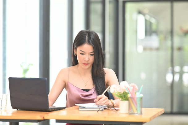 Una hermosa mujer asiática está sentada frente a un ordenador portátil en el escritorio de madera. - Foto, Imagen