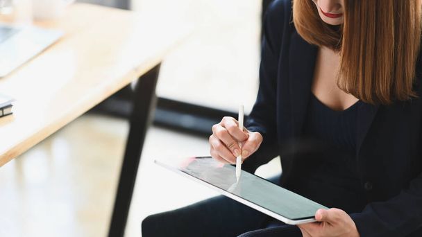 Zugeschnittene Aufnahme sexy Geschäftsfrau hält ein Computer-Tablet und Stift, während sie am hölzernen Arbeitstisch über dem gemütlichen Wohnzimmer als Hintergrund sitzt. - Foto, Bild