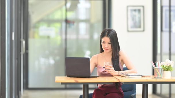 Güzel bir Asyalı kadın ahşap çalışma masasında bilgisayarın önünde oturuyor.. - Fotoğraf, Görsel