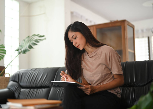 黒い革のソファに座っている間、女性はコンピュータタブレットとスタイラスペンを使用しています。. - 写真・画像