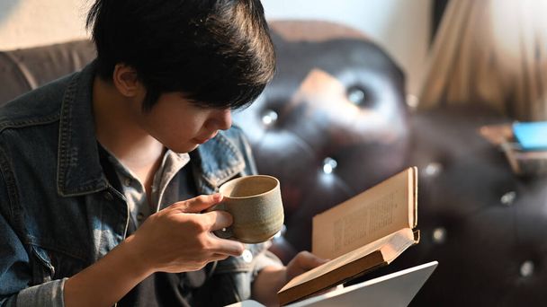 Mężczyzna z bliska czyta książkę i trzyma filiżankę kawy w dłoniach siedząc na skórzanej kanapie w wygodnym salonie jako tło. Mężczyzna odpoczywał podczas kwarantanny w domu.. - Zdjęcie, obraz