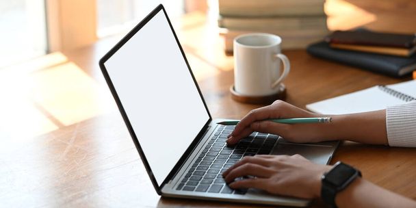 Oříznutý obrázek krásné ženy pomocí počítače notebook s bílou prázdnou obrazovkou, zatímco sedí na bílém pracovním stole přes pohodlný obývací pokoj jako pozadí. - Fotografie, Obrázek