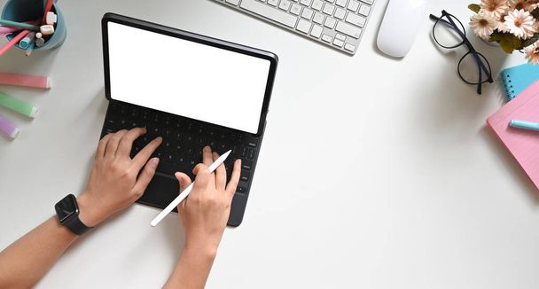 Τα χέρια πάνω όψης κρατούν ένα στυλό και πληκτρολογούν σε μια λευκή κενή οθόνη tablet υπολογιστή στο λευκό χώρο εργασίας. - Φωτογραφία, εικόνα