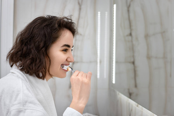 woman in white bathrobes brushing teeth in front of mirror - Foto, afbeelding